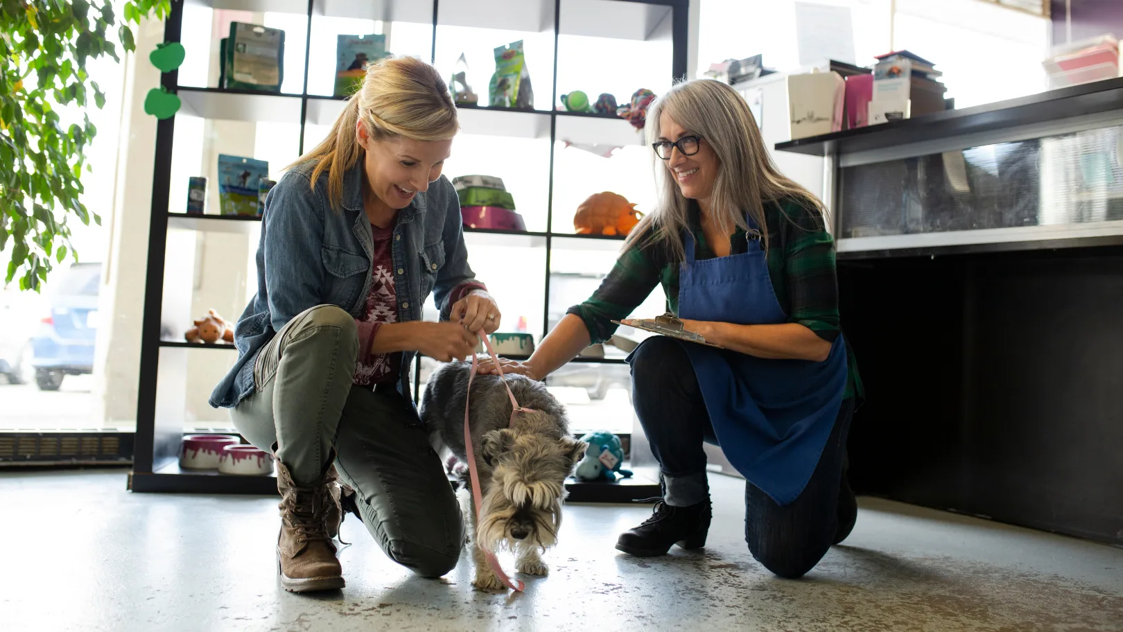 Two women crouch on the floor of a pet store with a small gray and white dog
