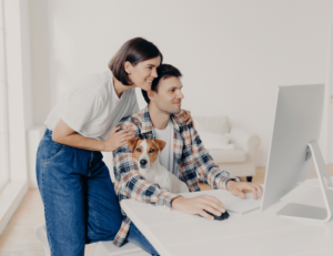 A woman and a man sit at a desk with a computer, discussing automated reminders, as a small dog curls up on the man's lap.