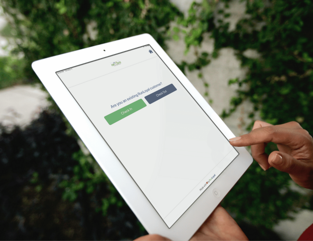 A person is using a white tablet with the RunLoyal interface for check-ins and check-outs, set against a backdrop of vibrant greenery. This seamless Dog Business Software simplifies management for daycare or grooming & more services.