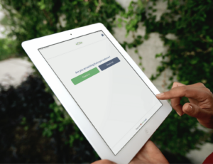 A person is using a white tablet with the RunLoyal interface for check-ins and check-outs, set against a backdrop of vibrant greenery. This seamless Dog Business Software simplifies management for daycare or grooming & more services.
