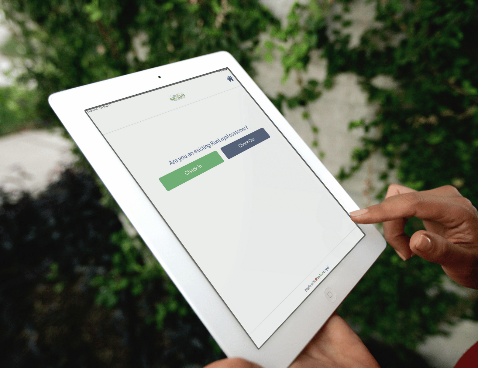 A person is using a white tablet with the RunLoyal interface for check-ins and check-outs, set against a backdrop of vibrant greenery. This seamless Dog Business Software simplifies management for daycare or grooming & more services.