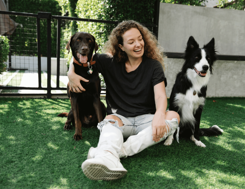 Woman sitting on grass with an arm around a brown dog, while a black and white dog sits nearby, all in an enclosed yard.