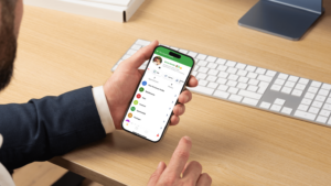 A person in a suit holds a smartphone displaying a messaging app interface at a desk with a computer keyboard and monitor, perhaps discussing ideas for the Scent Room.