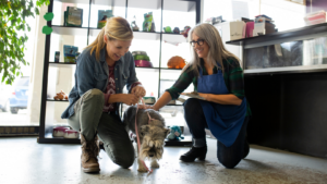 Two women crouch on the floor of a pet store, interacting with a small gray and white dog between them. Shelves of pet products and vaccination tracking materials are visible in the background.